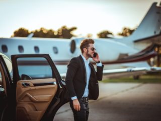 Young rich businessman talking on a mobile phone while getting out of a luxurious car parked next to a private airplane on a tarmac. - Journey House Travel